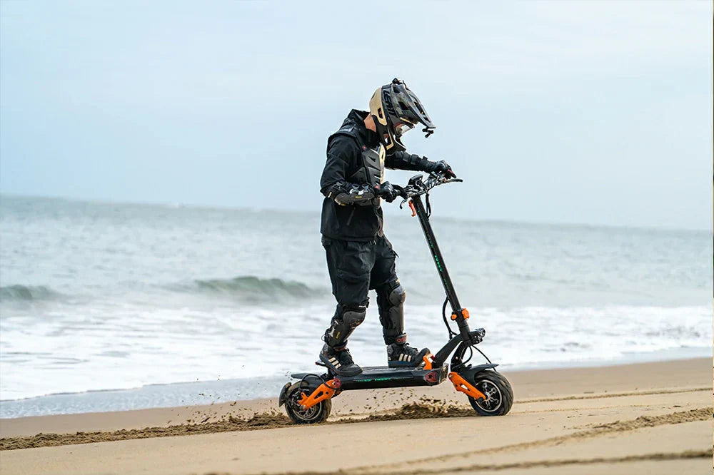 Dynamic seaside action shot of a rider in full protective gear driving black-orange Freeboy H9 electric scooter along sandy beach shore beside gentle ocean surf.