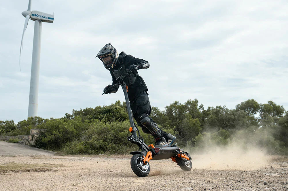Rider in full protective gear drifting on Freeboy H9 black‑orange off‑road electric scooter on gravel terrain with wind turbine and green shrubs in background
